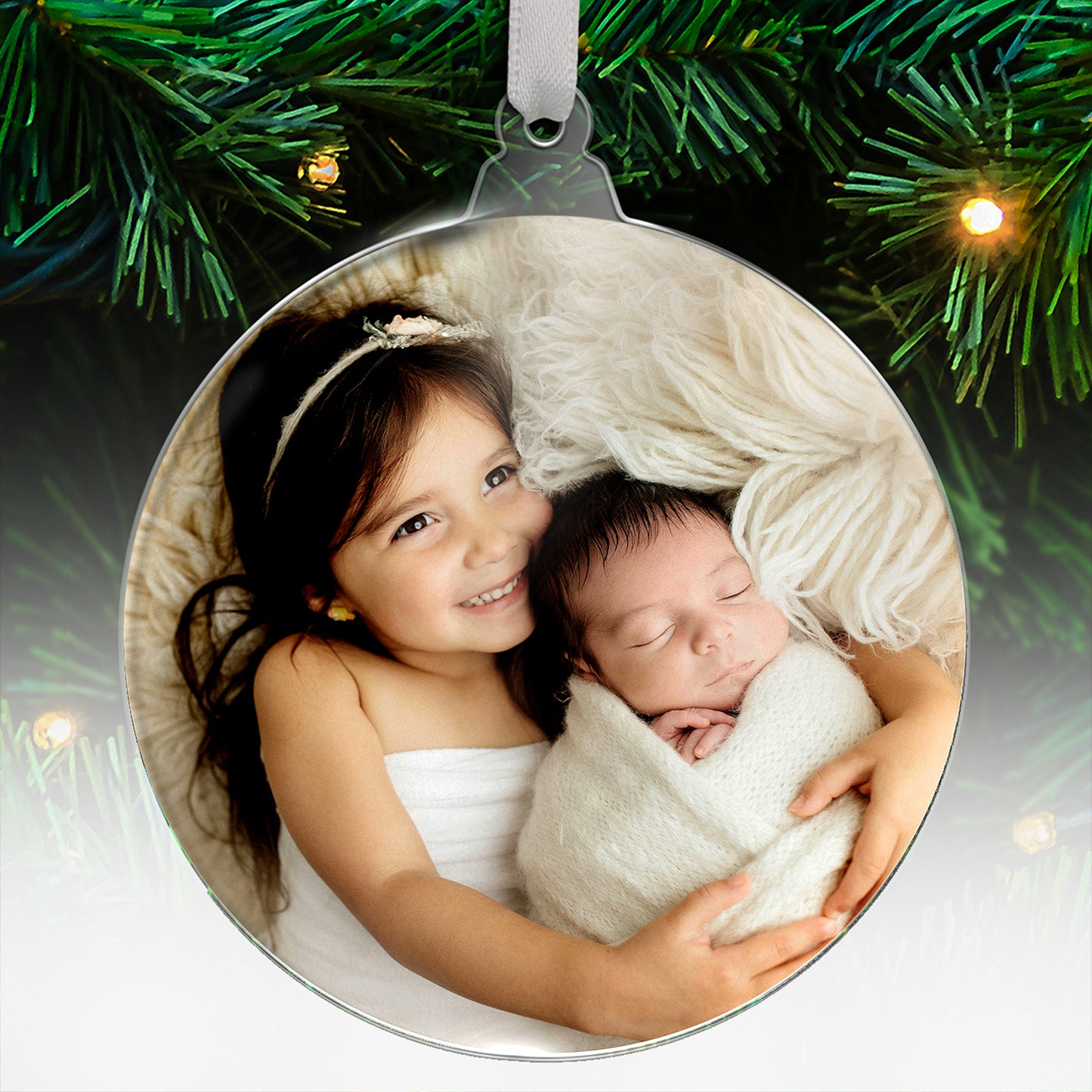Personalised acrylic Christmas tree ornament featuring an uploaded photograph of a young girl cuddling a sleeping baby, with festive evergreen branches and lights in the background.