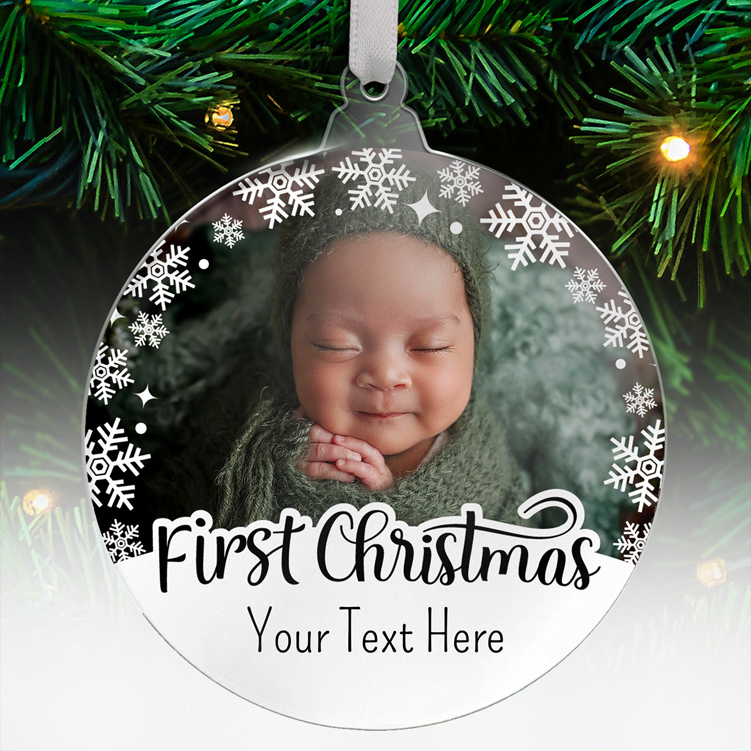 Round acrylic Christmas tree ornament with a photo uloaded of a baby and decorative white snowflakes around the border, featuring the words 'First Christmas' and below 'Your Text Here' in a tall font, hanging on a tree with green branches and fairy lights.