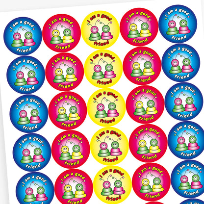 A sheet of round reward stickers in various bright colours, each with the phrase 'I am a good friend' written in bold, playful text. Each sticker features two cartoon alien-like characters with antennae, sitting side by side and smiling. The background colours include red, blue, yellow, and pink gradients, creating a cheerful and encouraging design aimed at promoting positive social behaviour.
