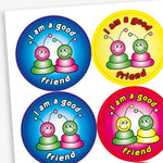 A sheet of round reward stickers in various bright colours, each with the phrase 'I am a good friend' written in bold, playful text. Each sticker features two cartoon alien-like characters with antennae, sitting side by side and smiling. The background colours include red, blue, yellow, and pink gradients, creating a cheerful and encouraging design aimed at promoting positive social behaviour.