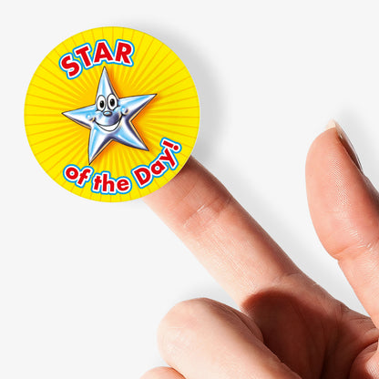 Star of the Day' reward stickers featuring smiling cartoon star with expressive face. Yellow background. Shown on a finger.