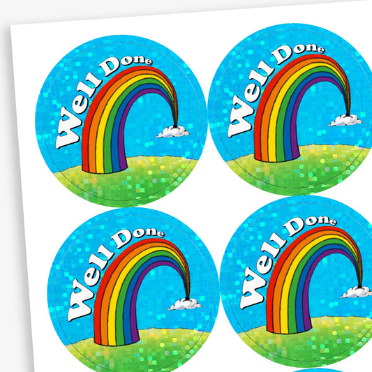 A sheet of holographic circular reward stickers. Each sticker features a colourful rainbow arcing from grassy hills toward small clouds, set against a bright blue sky with sparkly accents. The words 'Well Done' are written in bold white text along the curve of the rainbow.