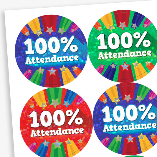 A sheet of colourful circular stickers with the text '100% Attendance' in bold white letters. Each sticker features a vibrant holographic background with rainbow-coloured beams and stars, in various base colours including blue, green, red, and purple.