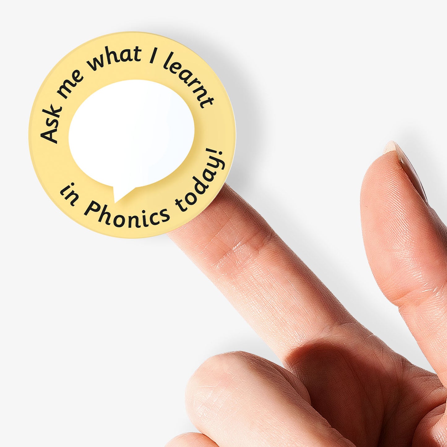 Circular sticker with text 'Ask me what I learnt in Phonics today!' held by a finger on a white background