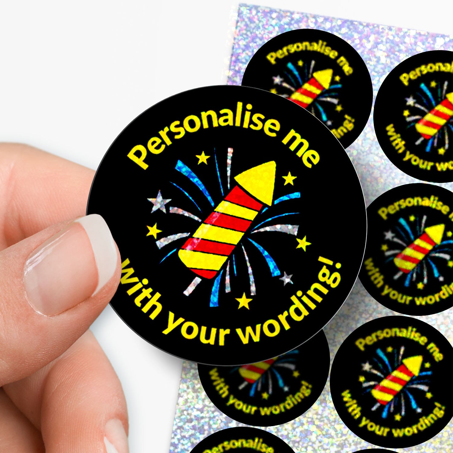 Circular sticker with sparkly firework design and personalised text held by hand with sticker sheet in the background.