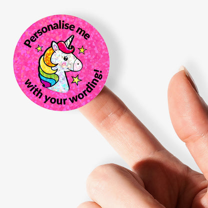 Pink round sticker with a unicorn design and text 'Personalise me with your wording!' held between fingers.