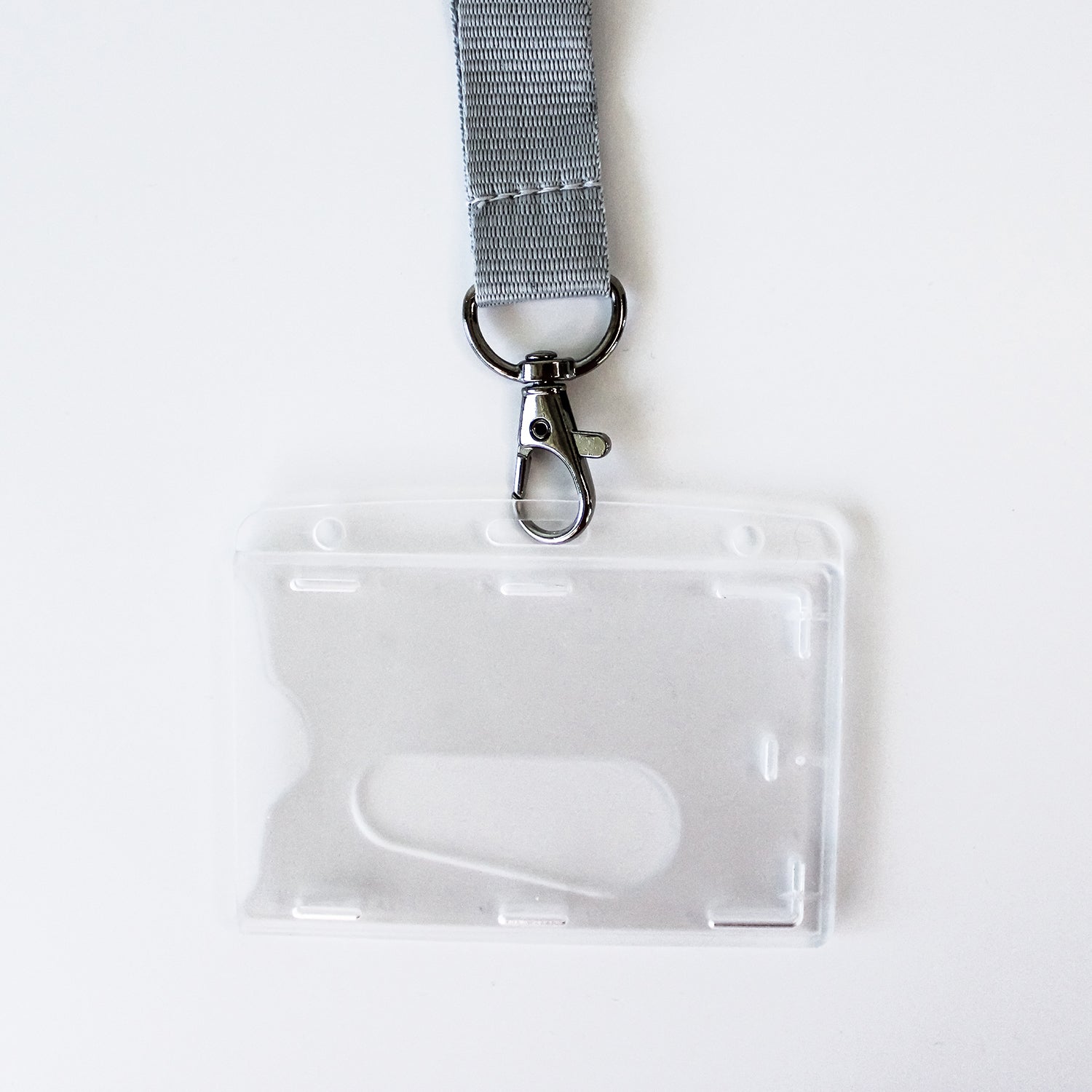 A grey woven lanyard with a silver metal clasp attached to a clear plastic ID badge holder, photographed on a plain white background.