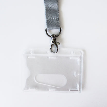 A grey woven lanyard with a silver metal clasp attached to a clear plastic ID badge holder, photographed on a plain white background.