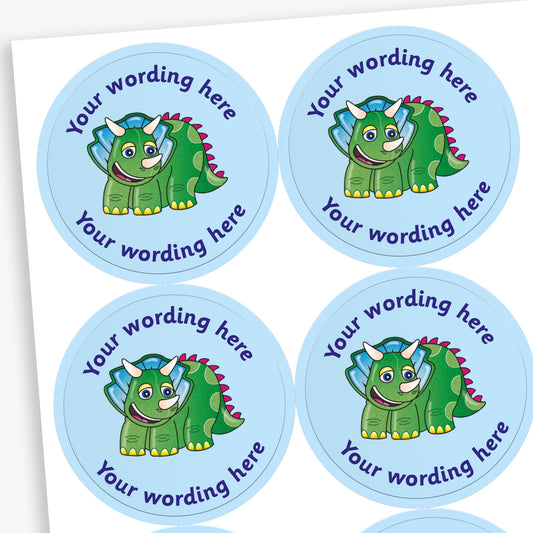 A sheet of pale blue circular stickers featuring a smiling cartoon Triceratops dinosaur; curving above and below the image is space for personalisation 'Your wording here' in a clear, bold font.