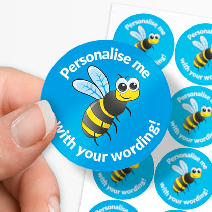 Hand holding a blue sticker with a bee design and text 'Personalise me with your wording!' against a white background.