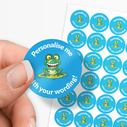 Circular blue sticker with a green frog graphic and text 'Personalise me with your wording!' held by a hand.