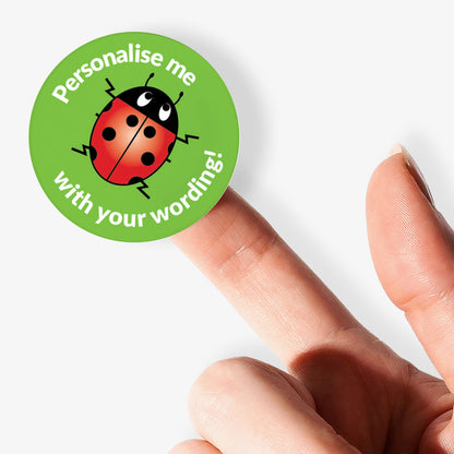 Round sticker with a ladybug design and text 'Personalise me with your wording!' held between fingers.