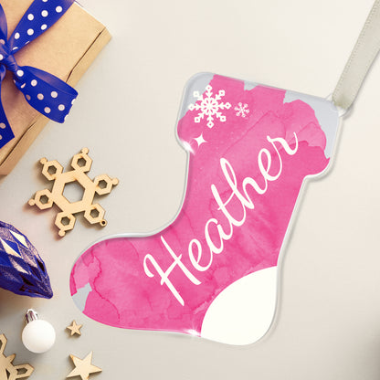 Personalised Christmas stocking-shaped acrylic decoration with the name 'Heather' written in white script on a pink background, featuring a snowflake design, placed on a white surface beside a present and christmas decorations.