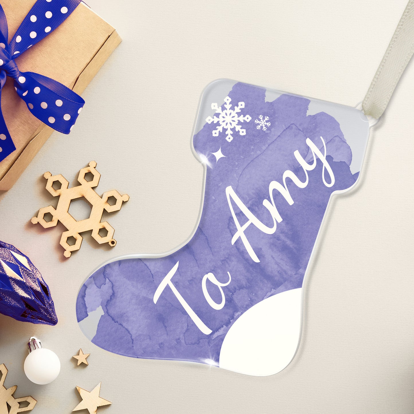 Personalised Christmas stocking-shaped acrylic decoration / gift tag with the name 'To Amy' written in white script on a watercolour blue background, featuring a snowflake design, placed on a white surface beside a present and christmas decorations.