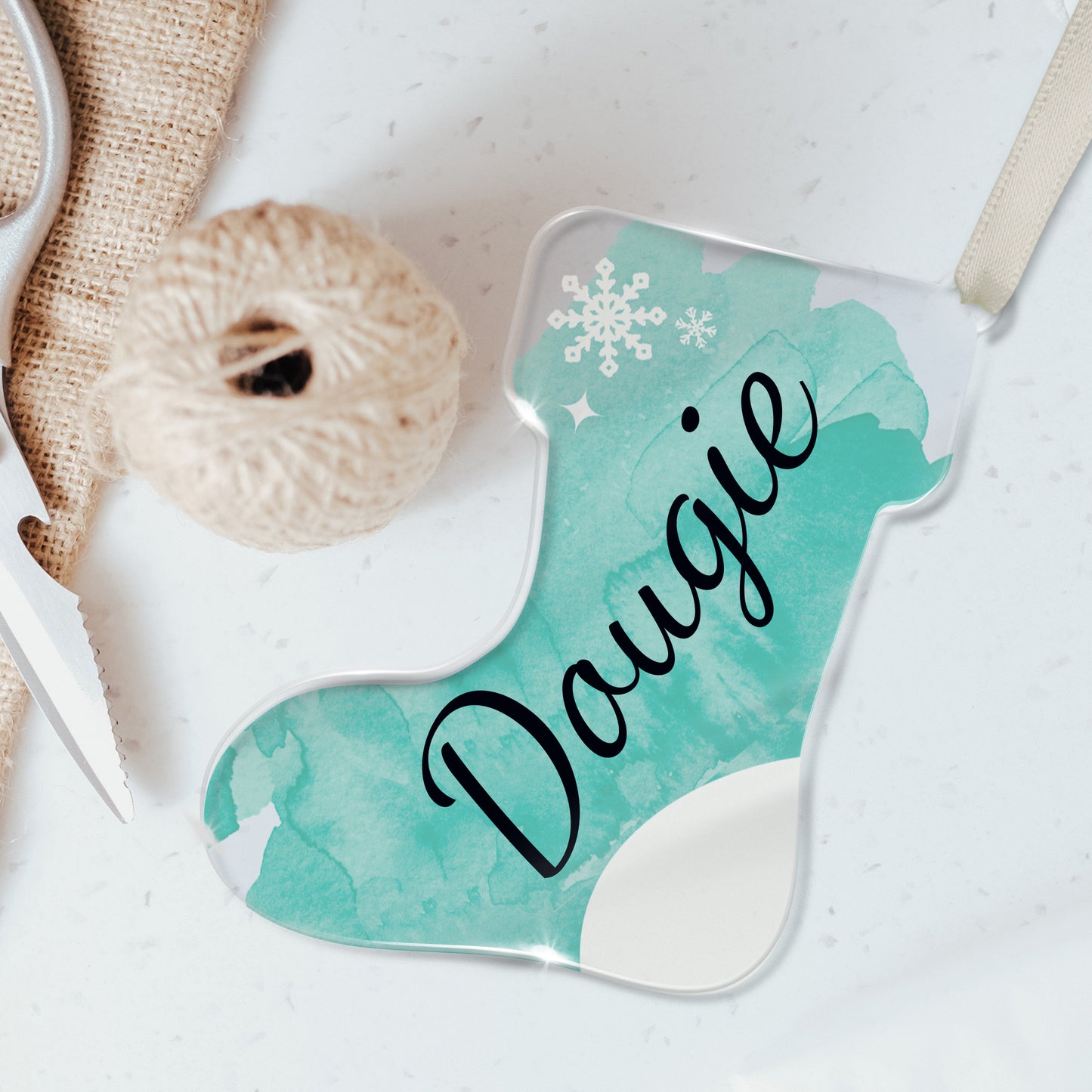 Personalised Christmas stocking-shaped acrylic decoration with the name 'Dougie' written in black script on a cyan background, featuring a snowflake design, placed on a white surface beside twine and scissors