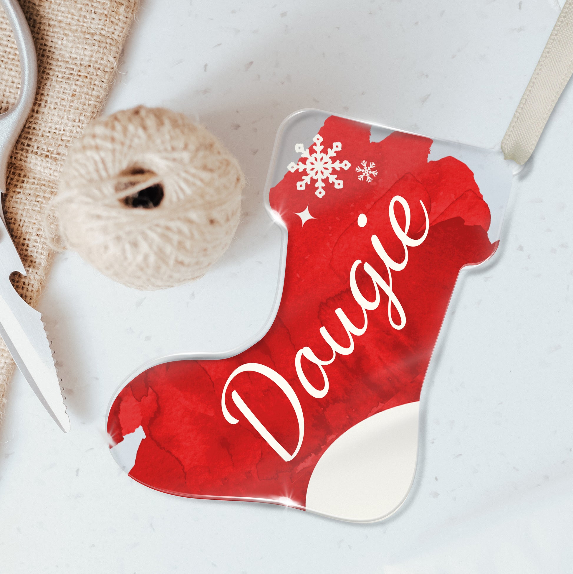 Personalised Christmas stocking-shaped acrylic decoration with the name 'Dougie' written in white script on a red background, featuring a snowflake design, placed on a white surface beside twine and scissors