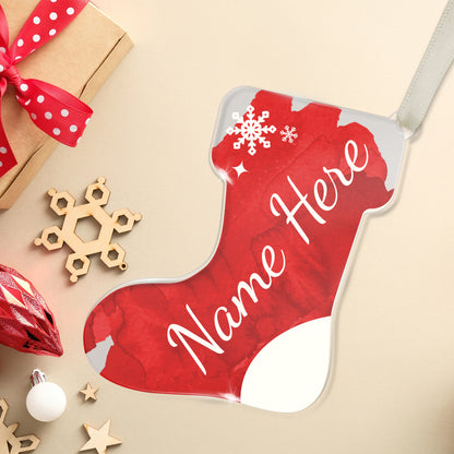 Personalised Christmas stocking-shaped acrylic decoration with the customisable place holder 'Name Here' written in white script on a red background, featuring a snowflake design, placed on a white surface beside a present and christmas decorations.