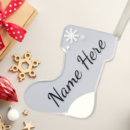 Personalised Christmas stocking-shaped acrylic decoration with the customisable place holder 'Name Here' written in black script on a clear background, featuring a snowflake design, placed on a white surface beside a present and christmas decorations.