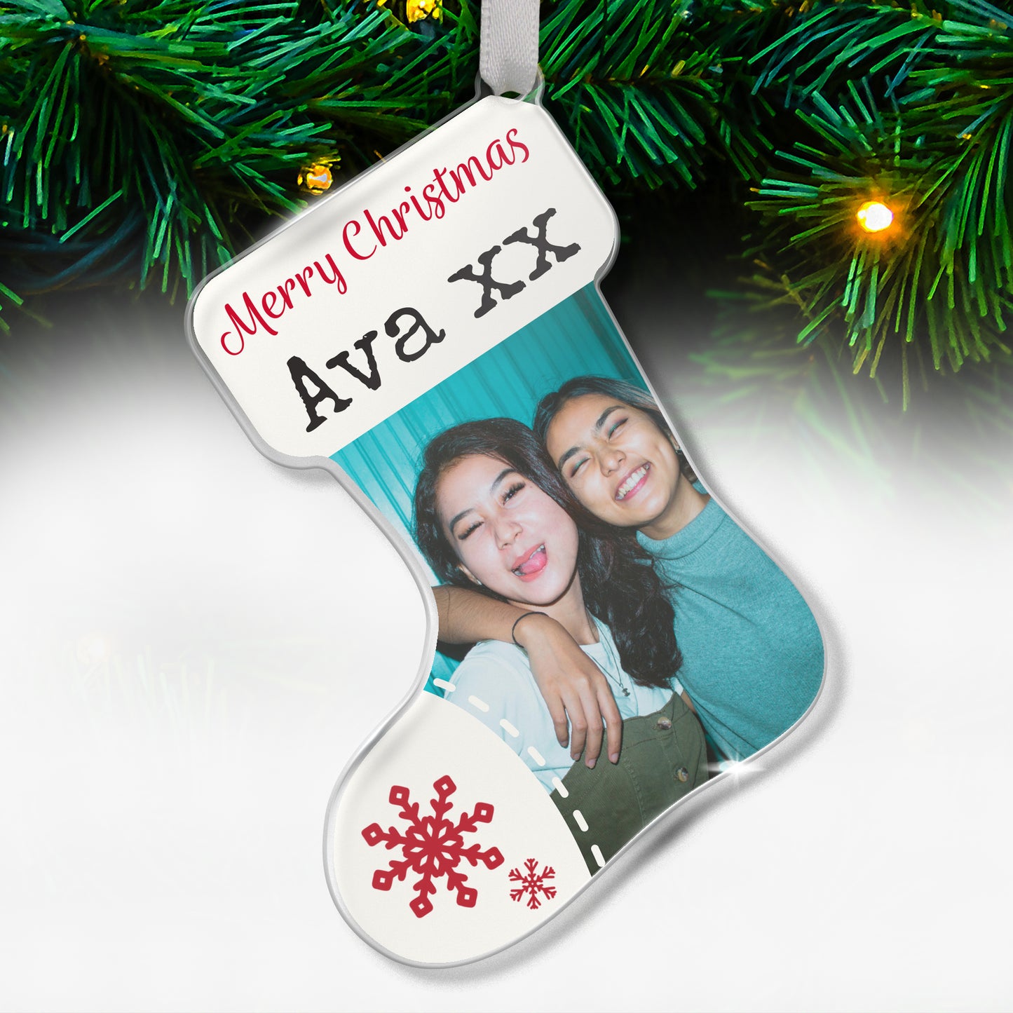 Stocking-shaped acrylic Christmas tree ornament with a white design, red snowflake decorations, and the message 'Merry Christmas Ava XX' at the top, featuring a photo of two smiling friends hugging each other, hanging on a tree with fairy lights.