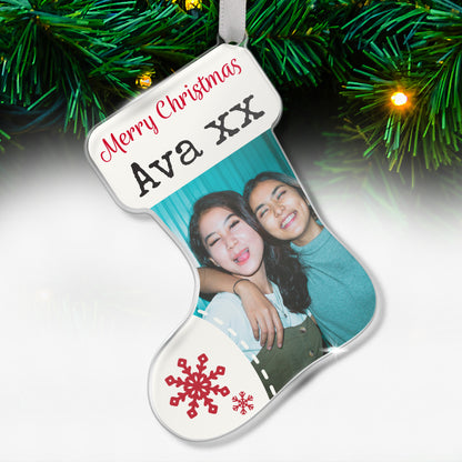 Stocking-shaped acrylic Christmas tree ornament with a white design, red snowflake decorations, and the message 'Merry Christmas Ava XX' at the top, featuring a photo of two smiling friends hugging each other, hanging on a tree with fairy lights.