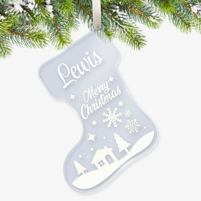 Clear acrylic Christmas stocking-shaped tree ornament decorated with a white winterscene of snow falling on a house with trees, featuring a space at the top for a customisation saying 'Lewis' in white script, with further script writing below in white saying 'Merry Christmas' hanging on a Christmas tree.