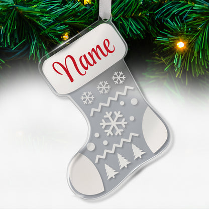 Clear acrylic Christmas stocking-shaped tree ornament decorated with white snowflakes, zigzags, and tree patterns, featuring a space at the top for a personalised name in red script, hanging on a Christmas tree with lights.