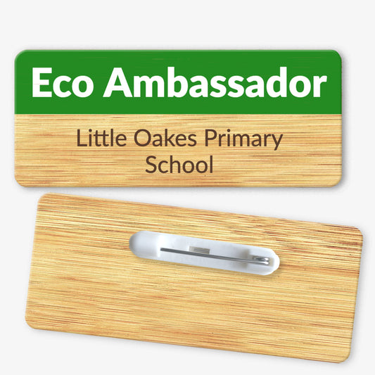 A rectangular bamboo name badge with a natural background and a green top section. The text reads 'Eco Ambassador' in bold white letters on the green part, and 'Little Oakes Primary School' in black below. The reverse side of the badge is also shown, featuring a metal safety pin fastening for attachment.