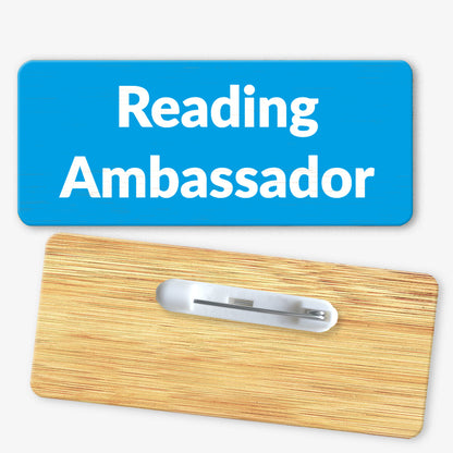 A rectangular bamboo name badge with a cyan background. The text reads 'Reading Ambassador' in bold white letters. The reverse side of the badge is also shown, featuring a metal safety pin fastening for attachment.