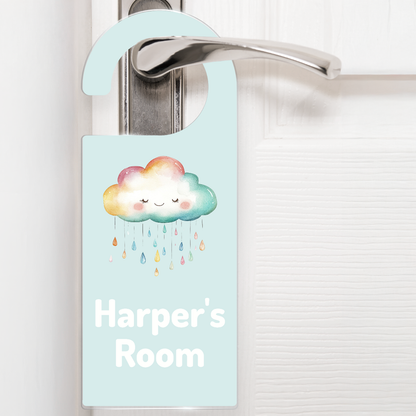 A light green door hanger hanging on a silver handle, featuring a cute rainbow cloud with a smiling face and raining rainbow drops above the text 'Harper's Room' in white letters. The design is playful and child-friendly, ideal for decorating a child's bedroom door.