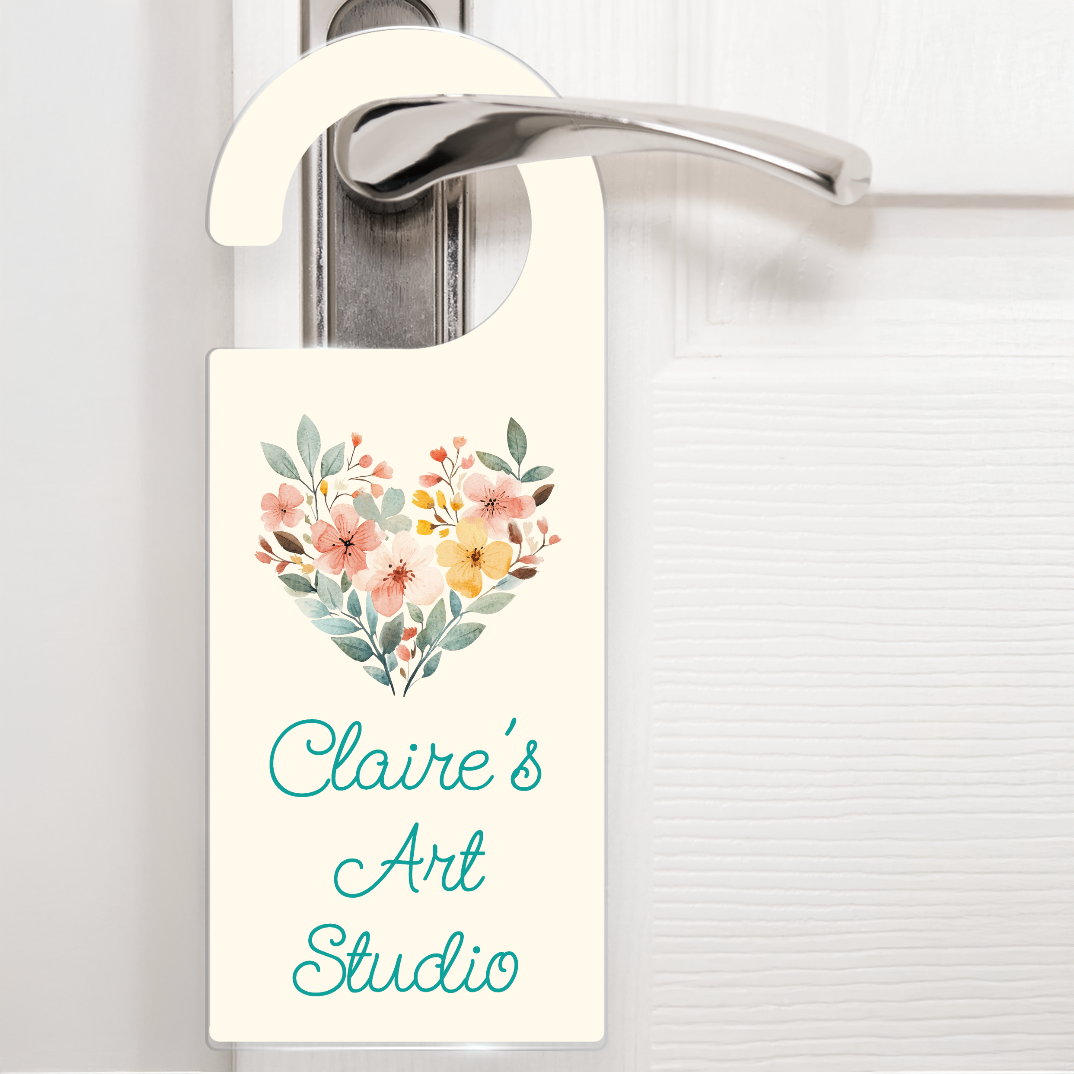 A cream door hanger hanging on a silver handle, featuring a heart mades of watercolour flowers above the text 'Claire's Art Studio' in aqua script writing. The design is sylish and delicate - ideal for identifying different rooms.