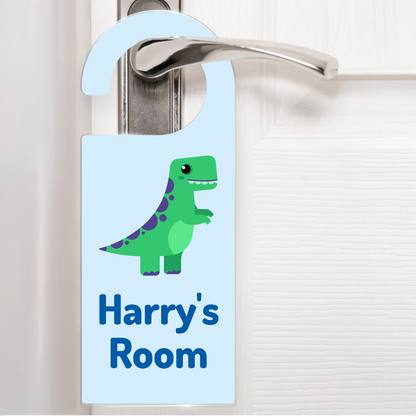 A pale blue door hanger hanging on a silver handle, featuring a cartoon green and purple dinosaur above the text 'Harry's Room' in darker blue letters. The design is playful and child-friendly, ideal for decorating a child's bedroom door.