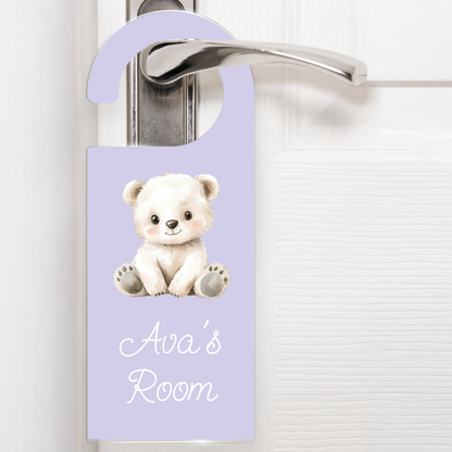 A lilac door hanger hanging on a silver handle, featuring a cute cartoon white bear above the text 'Ava's Room' in white letters. The design is playful and child-friendly, ideal for decorating a child's bedroom door.