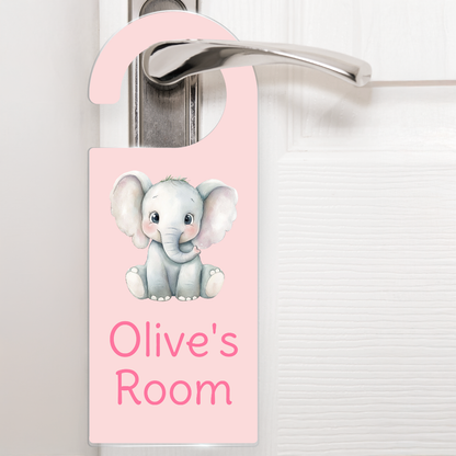 A pale pink door hanger hanging on a silver handle, featuring a cute cartoon elephant above the text 'Olive's Room' in darker pink letters. The design is playful and child-friendly, ideal for decorating a child's bedroom door.