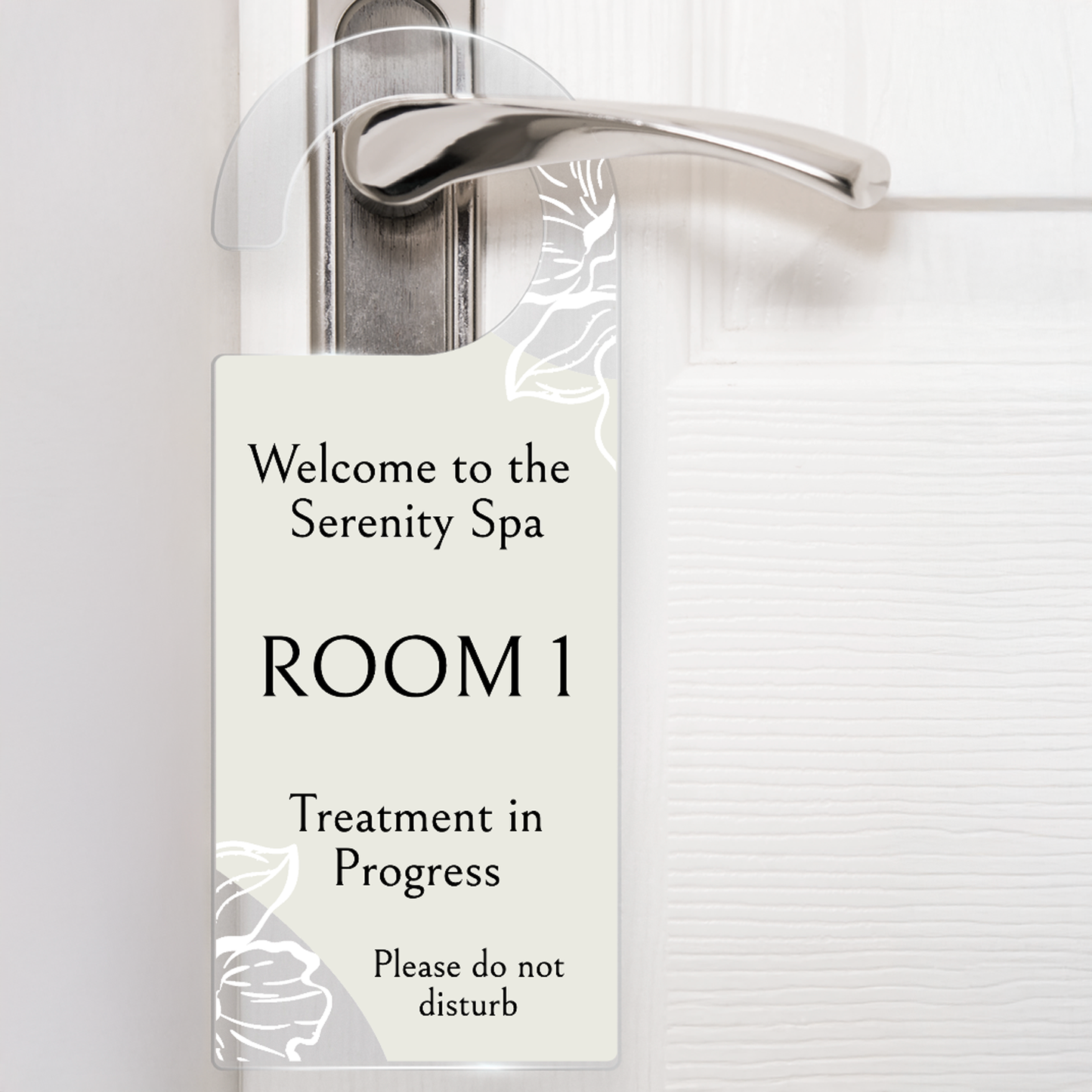 Acrylic door hanger suspened from a silver door handle against a white door.  The top wording says 'Welcome to the Serenity Spa' middle wording 'ROOM 1' Lower wording 'Treatment in Progress' and then bottom right 'Please do not disturb'.  The door hanger has an off white background with delicate flowers at the handle and the bottom left corner.