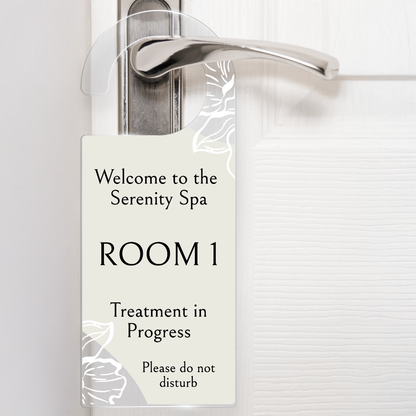 Acrylic door hanger suspened from a silver door handle against a white door.  The top wording says 'Welcome to the Serenity Spa' middle wording 'ROOM 1' Lower wording 'Treatment in Progress' and then bottom right 'Please do not disturb'.  The door hanger has an off white background with delicate flowers at the handle and the bottom left corner.
