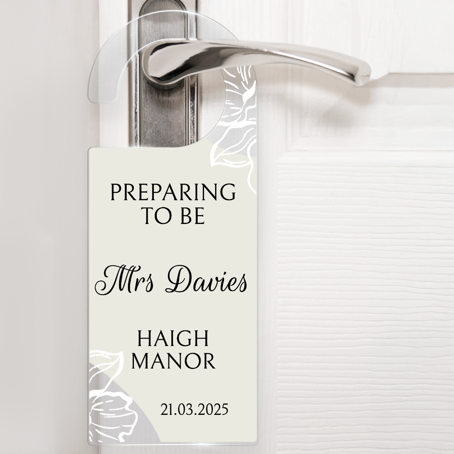 Acrylic door hanger suspened from a silver door handle against a white door.  The top wording says 'PREPARING TO BE' middle wording 'Mrs Davies' Lower wording 'HAIGH MANOR' and then bottom right '21.03.2025'.  The door hanger has an off white background with delicate flowers at the handle and the bottom left corner.