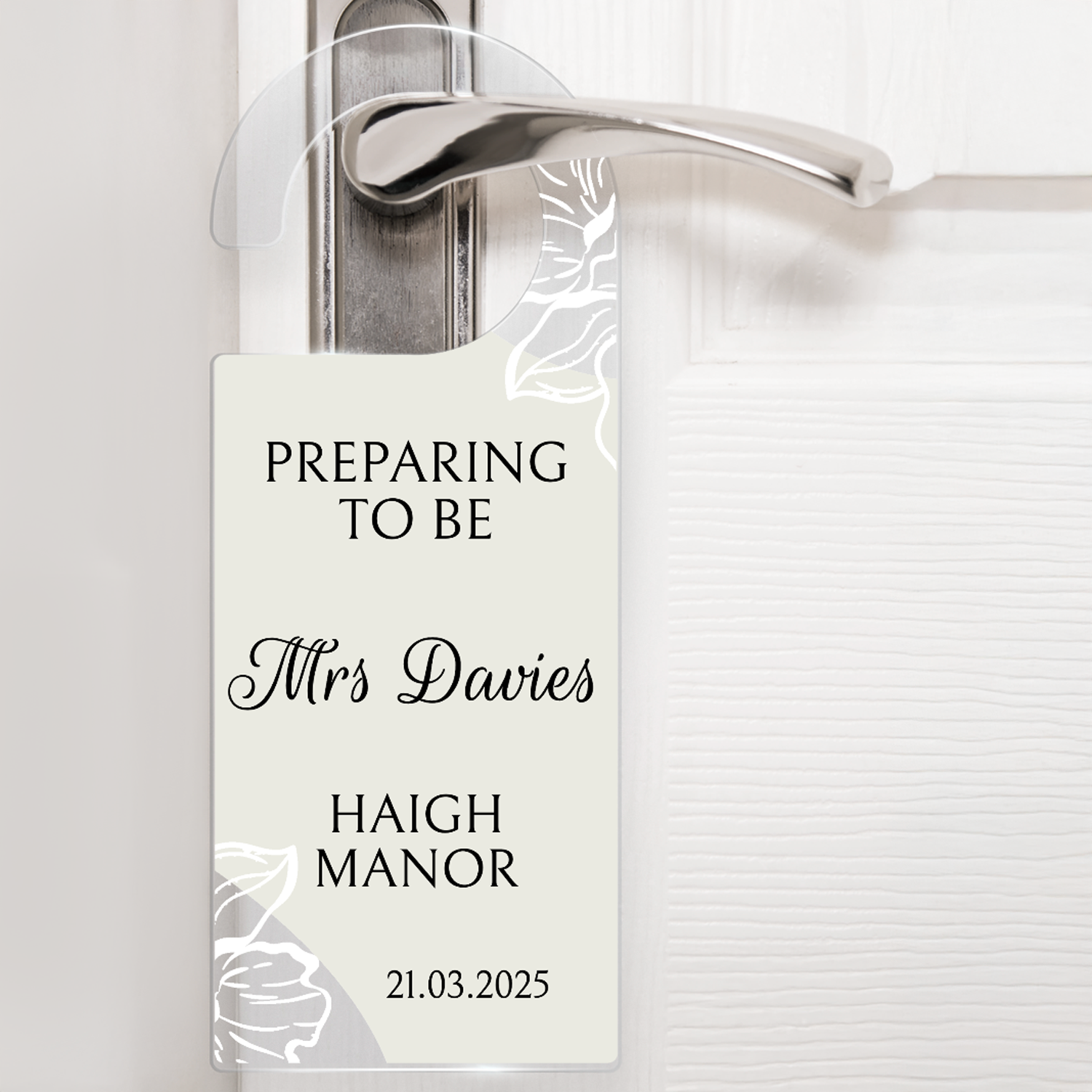Acrylic door hanger suspened from a silver door handle against a white door.  The top wording says 'PREPARING TO BE' middle wording 'Mrs Davies' Lower wording 'HAIGH MANOR' and then bottom right '21.03.2025'.  The door hanger has an off white background with delicate flowers at the handle and the bottom left corner.