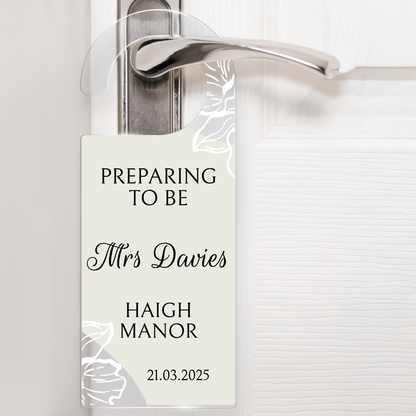 Acrylic door hanger suspened from a silver door handle against a white door.  The top wording says 'PREPARING TO BE' middle wording 'Mrs Davies' Lower wording 'HAIGH MANOR' and then bottom right '21.03.2025'.  The door hanger has an off white background with delicate flowers at the handle and the bottom left corner.