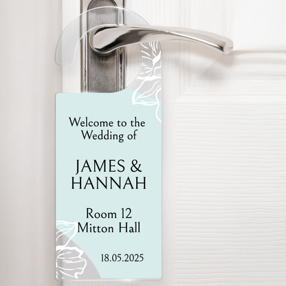 Acrylic door hanger suspened from a silver door handle against a white door.  The top wording says 'Welcome to the Wedding of' middle wording 'JAMES & HANNAH' Lower wording 'Room 12 Mitton Hall' and then bottom right '18.05.2025'.  The door hanger has an pale blue background with delicate flowers at the handle and the bottom left corner.