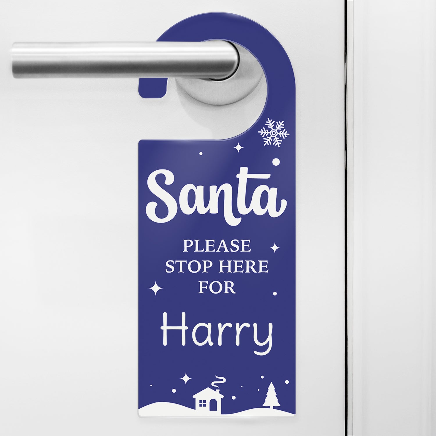 A festive dakr blue door hanger with white text that reads 'Santa PLEASE STOP HERE FOR Harry'. The design includes snowflakes, stars, and a snowy village scene with a house and trees at the bottom. The hanger is displayed on a silver door handle, ready for Christmas Eve.
