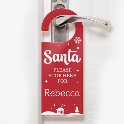 A festive red door hanger with white text that reads 'Santa PLEASE STOP HERE FOR Rebecca'. The design includes snowflakes, stars, and a snowy village scene with a house and trees at the bottom. The hanger is displayed on a silver door handle, ready for Christmas Eve.
