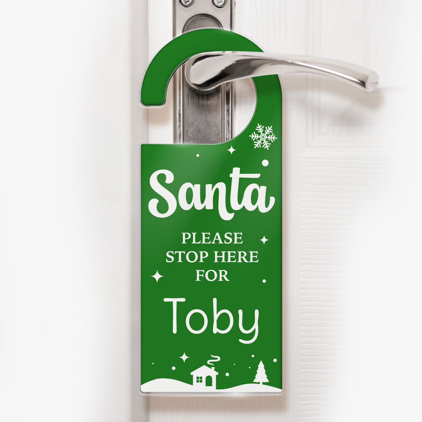 A festive green door hanger with white text that reads 'Santa PLEASE STOP HERE FOR Toby'. The design includes snowflakes, stars, and a snowy village scene with a house and trees at the bottom. The hanger is displayed on a silver door handle, ready for Christmas Eve.
