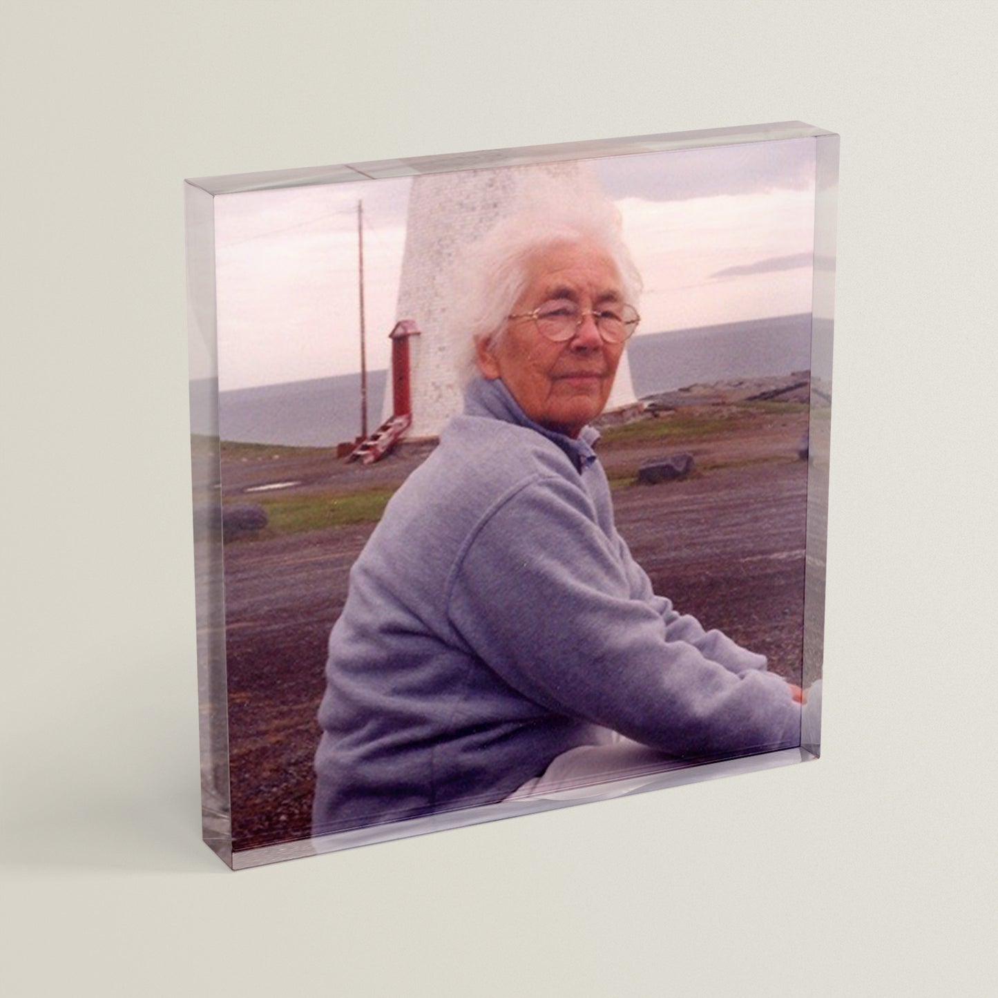 A square acrylic photo block featuring a photo of an older lady in front of a lighthouse.  The block has a glossy, freestanding design ideal for display.