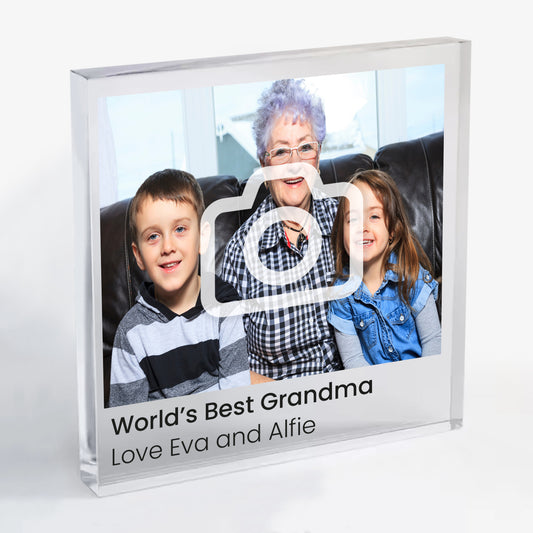 A square acrylic photo block featuring a family photo. The image shows an elderly woman sitting on a sofa with two smiling children, one on each side. Beneath the photo, printed text reads 'World’s Best Grandma' followed by 'Love Eva and Alfie'. The block has a glossy, freestanding design ideal for display.