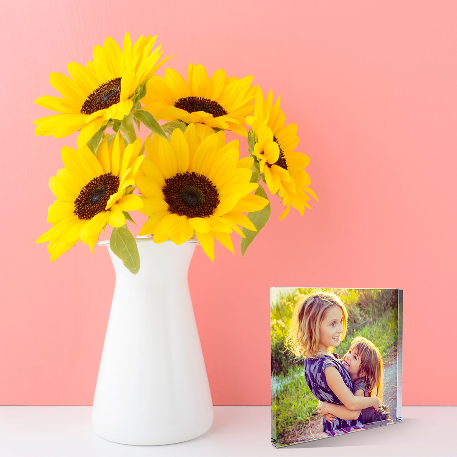 A square acrylic photo block featuring a family photo. The image shows the full areas printed with the photo of two smiling children.  The block has a glossy, freestanding design ideal for display.  The photo bloack is stood next to a vase of sunflowers.