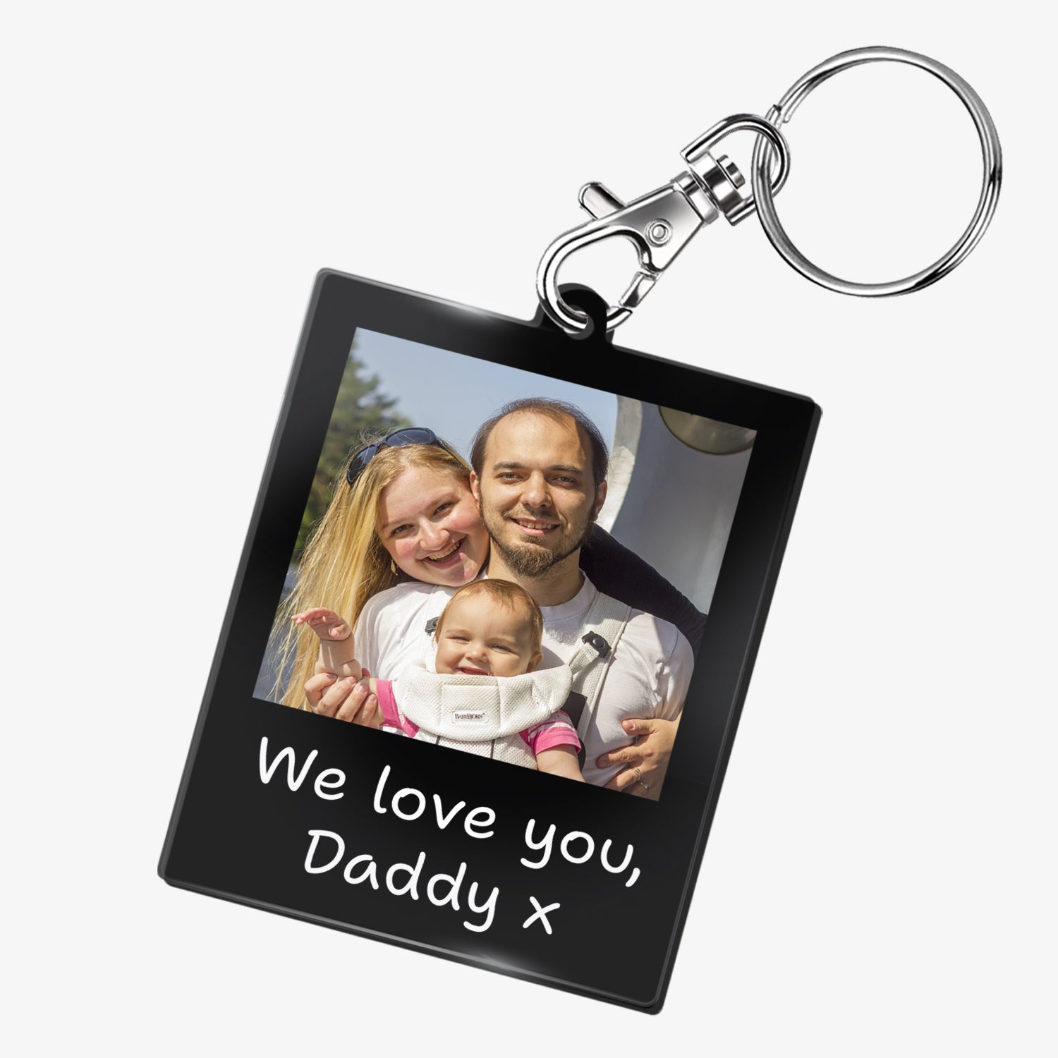 A personalised keyring featuring a rectangular photo frame design with a metal keychain and clasp. The photo shows a family. Below the image, there is placeholder text reading 'We love you, Daddy x'. The keyring has a black glossy finish and a polaroid-style layout.