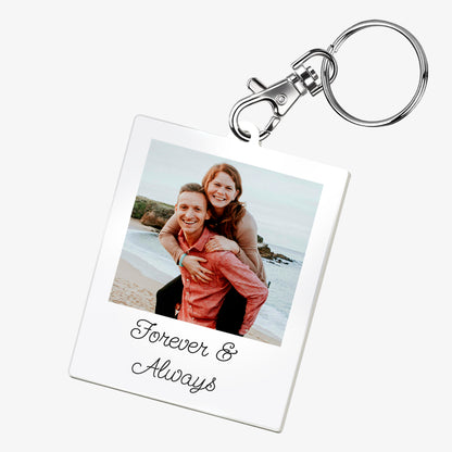 A personalised keyring featuring a rectangular photo frame design with a metal keychain and clasp. The photo shows a couple on a beach. Below the image, there is placeholder text reading 'Forever & Always'. The keyring has a glossy finish and a polaroid-style layout.