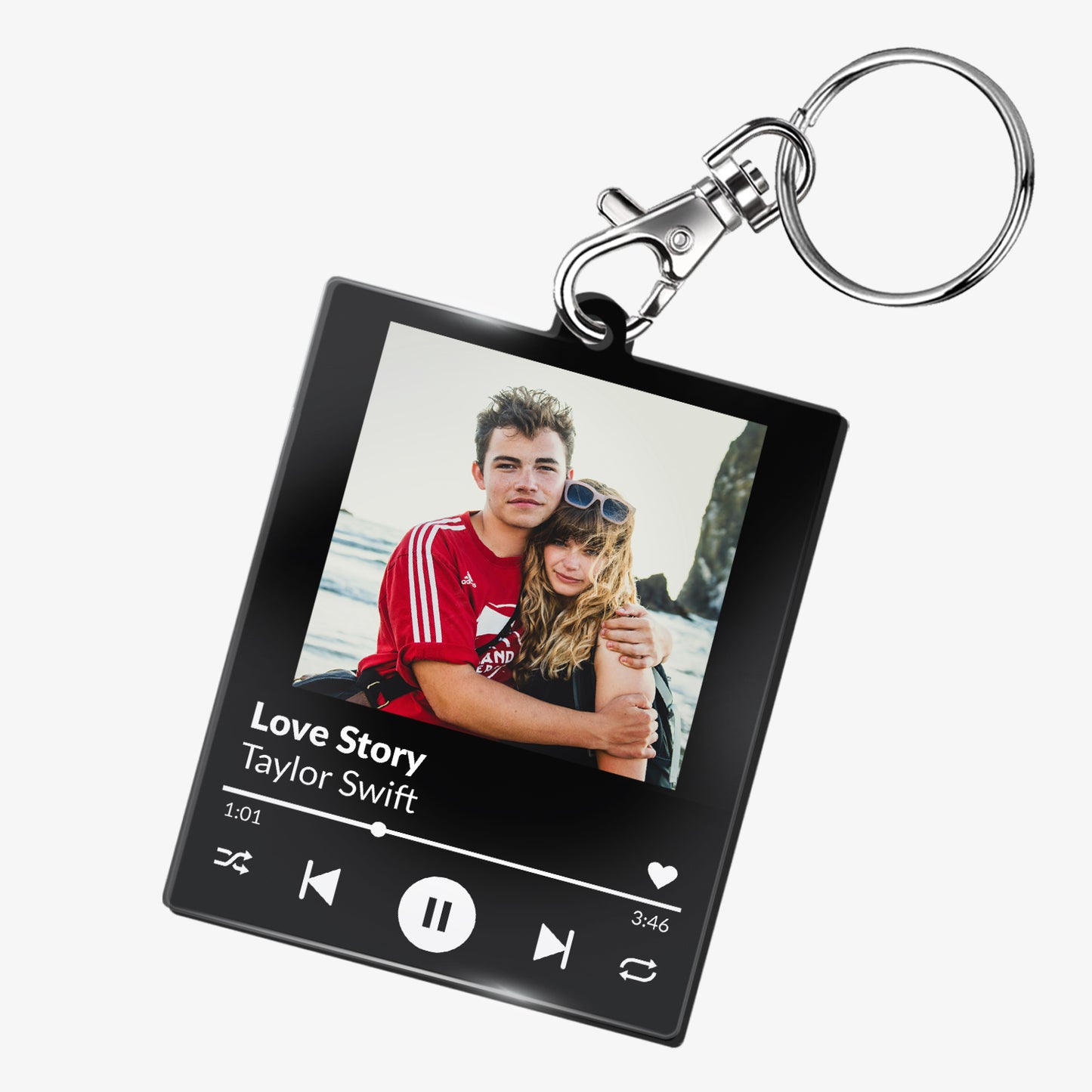 A custom acrylic keyring designed to resemble a digital music player interface. It features a square frame with a photo of two smiling people at the top. Below the photo are mock song details, including placeholder text 'Song Title Here' and 'Artist Name'. Music control icons—rewind, play, forward, and a heart—are displayed along the bottom, mimicking a streaming app. The keyring is attached to a metal clasp and keyring loop.
