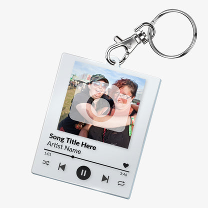 A custom acrylic keyring designed to resemble a digital music player interface. It features a square frame with a photo of two smiling people at the top. Below the photo are mock song details, including placeholder text 'Song Title Here' and 'Artist Name'. Music control icons—rewind, play, forward, and a heart—are displayed along the bottom, mimicking a streaming app. The keyring is attached to a metal clasp and keyring loop.