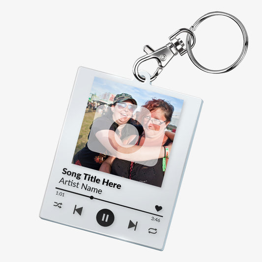 A custom acrylic keyring designed to resemble a digital music player interface. It features a square frame with a photo of two smiling people at the top. Below the photo are mock song details, including placeholder text 'Song Title Here' and 'Artist Name'. Music control icons—rewind, play, forward, and a heart—are displayed along the bottom, mimicking a streaming app. The keyring is attached to a metal clasp and keyring loop.